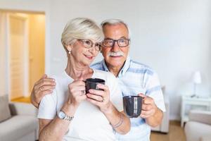 heureux couple de personnes âgées debout dans le salon. se regardant en souriant et tenant une tasse de café ou de thé. mari âgé étreignant sa charmante épouse photo