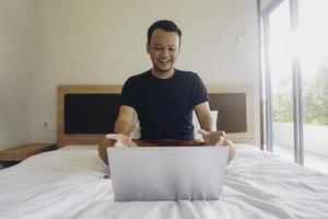 heureux jeune homme travaillant depuis le lit. placez l'ordinateur portable sur le lit et tapez sur le clavier. photo