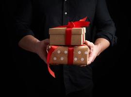 adulte homme dans une noir chemise détient une empiler de cadeaux enveloppé dans marron papier avec une rouge arc sur une noir Contexte photo