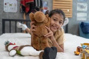 douce petite fille étreint un ours en peluche, regarde la caméra et sourit assise sur son lit à la maison. photo