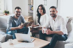 réussi équipe. Trois sur de soi affaires gens dans intelligent décontractée porter à la recherche à caméra et souriant tandis que séance ensemble à le bureau dans Bureau photo