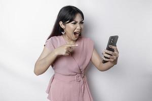 une mécontent Jeune asiatique femme habillé dans rose, regards mécontent avec irrité visage expressions en portant sa téléphone photo