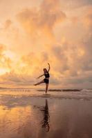 une ballerine dans une noir robe est pratiquant ballet se déplace sur le plage avec très souple mouvements avec une vue de le des nuages derrière photo