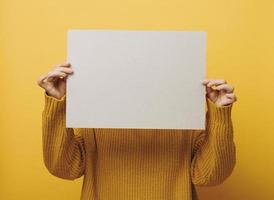 femme dans un chandail orange tient une feuille de papier vierge sur fond jaune. place pour une inscription, une publicité, une information photo