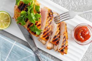 Haut vue de tranches de tranché grillé poulet filet Sein sur une portion blanc rectangulaire assiette avec sauce et chaux. grand espèces. fourchette et couteau. photo