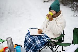 femme asseoir dans chaise, lis livre et boisson thé dans hiver forêt sur une pique-nique. photo