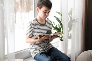 une six ans garçon est en train de lire une livre tandis que séance par le la fenêtre. Accueil scolarité photo