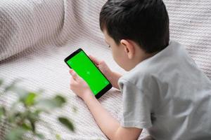 le garçon regards à le écran de le téléphone avec une chrome clé à l'intérieur. un méconnaissable enfant les usages une téléphone intelligent avec une vert écran à regarder une vidéo. photo