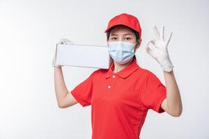 image d'un jeune livreur conscient en casquette rouge t-shirt blanc uniforme masque facial gants debout avec boîte en carton blanc vide isolé sur fond gris clair studio photo