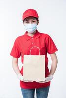 image d'un jeune livreur heureux en casquette rouge t-shirt blanc uniforme masque facial gants debout avec paquet de papier kraft marron vide isolé sur fond gris clair studio photo