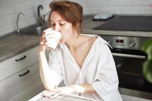 content magnifique femme avec tasse de café ou thé dans le blanc chemise séance sur cuisine dans le maison. photo