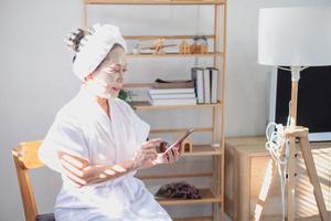 asiatique personnes âgées femme séance portant masque appliquant faciale crème mettre une serviette plus de votre tête portant une blanc manteau en train de lire carnet et mobile téléphone sur en bois chaise pour bien faciale peau santé photo