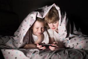 frère et sœur mensonge en dessous de une couverture à nuit et jouer sur le téléphone photo