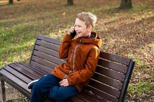 une garçon est parlant sur le téléphone et en riant tandis que séance sur une parc banc dans l'automne photo