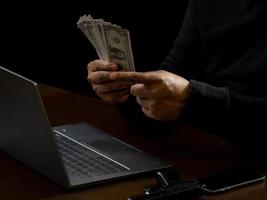 l'ordinateur et les mains d'un homme vêtu d'une chemise noire, assis sur une chaise et une table, est un voleur, détenant de l'argent, comptant le montant obtenu grâce au détournement ou au vol, dans une pièce noire. photo