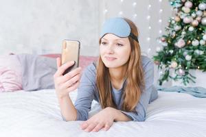 magnifique Jeune femme à la recherche à téléphone caméra et souriant tandis que mensonge dans lit photo