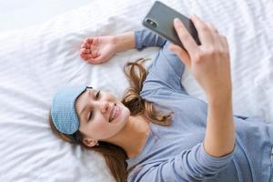 magnifique femme dans bleu pyjamas à la recherche dans téléphone. Matin départs avec téléphone. photo