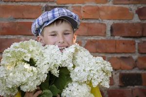 joyeux enfant de village heureux avec un bouquet de fleurs. petit garçon souriant avec hortensia près du mur de briques grunge. fête des mères, 8 mars, concept de la journée internationale de la femme. photo