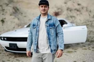 bel homme en veste de jeans et casquette marchant près de sa voiture de muscle blanche en carrière. photo