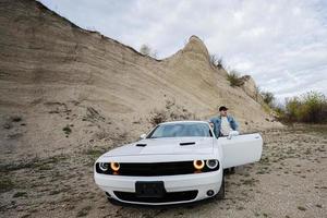 bel homme en veste de jeans et casquette se tient près de sa voiture de muscle blanche en carrière. photo