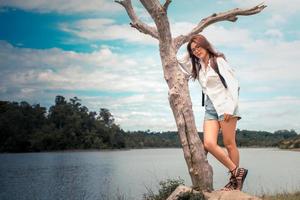 magnifique asiatique femme portant des lunettes et blanc chemise et jeans. permanent avec le sien droite main sur une mort arbre dans le Contexte est une vue de le rivière, montagnes, des nuages et ciel. à la recherche à caméra. photo