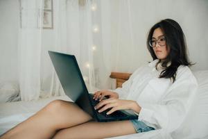 jolie attrayant asiatique femme portant des lunettes travail avec ordinateur portable, pose sur le blanc lit à chambre dans le matin, travail de Accueil La technologie femme ou alternative Bureau free-lance concept. photo