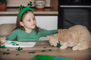 une peu fille avec une bandage sur sa tête dessine et coupes vert trèfles pour st. patrick's journée à une table à Accueil dans le cuisine, suivant à sa est sa magnifique chat avec une vert arc attacher autour le sien cou photo