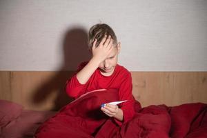 une mignonne garçon dans rouge pyjamas regards à le thermomètre et détient le sien tête avec le sien main photo