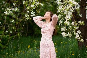 une fille dans une rose robe est permanent en dessous de une arbre avec sa yeux fermé et souriant photo