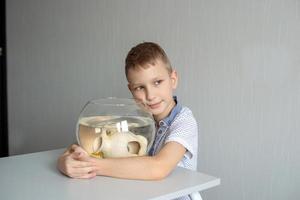 une mignonne garçon est assis près une transparent aquarium dans le pièce et câlins un aquarium avec poisson photo