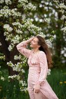 une fille dans une rose robe est permanent en dessous de une arbre avec sa yeux fermé et souriant photo