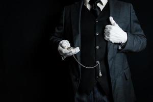 portrait de majordome ou serviteur dans foncé costume et blanc gants sur noir Contexte en portant poche montre. copie espace pour un service industrie et professionnel hospitalité. photo