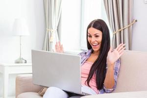 jolie jeune femme heureuse assise sur le canapé à l'aide d'un ordinateur portable, ayant un chat vidéo, agitant, parle avec son amie du coronavirus et reste à la maison pendant l'appel vidéo en ligne. travail à domicile photo