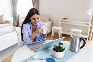 soi employé femme travail avec sa téléphone et portable à Accueil avec une tasse de café. content femme travail à Accueil avec portable. souriant femme en portant chaud tasse de thé photo