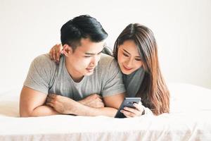 une Jeune asiatique couple pièces sur le téléphone dans lit dans le Matin dans le chambre à coucher. content famille concept photo