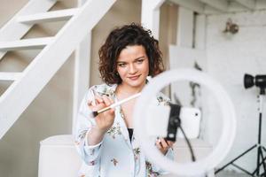Jeune brunette femme blogueur dans Accueil vêtements Faire maquillage avec mobile téléphone et bague lampe en ligne dans studio photo