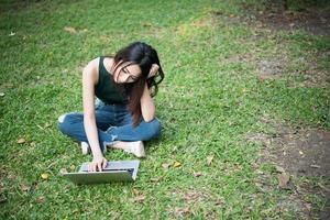 Belle jeune femme assise sur l'herbe verte et utilisant un ordinateur portable dans le parc photo