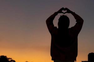 une silhouette de une Jeune femme élevage sa mains au dessus sa tête à représenter une cœur symbole signifiant amitié, l'amour et gentillesse. cœur symbole concept avec le sens de l'amour et amitié. photo