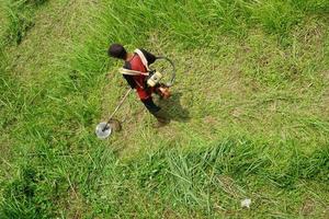 Haut vue de une homme dans une peignoir fauchage le herbe avec une tondeuse photo