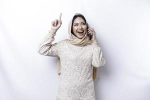 femme asiatique excitée portant le hijab pointant vers l'espace de copie au-dessus d'elle tout en tenant son téléphone, isolée sur fond blanc photo