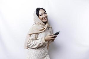 une portrait de une content asiatique musulman femme portant une hijab, en portant sa téléphone, isolé par blanc Contexte photo