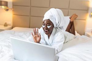 magnifique Jeune africain femme dans peignoir de bain repos sur lit après douche vidéo appel sa copains sur sa portable dans le matin.à la recherche à portable ordinateur et parlant à sa copains via vidéo appel. photo
