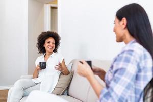 deux femelle copains dans social distanciation séance sur canapé dans coronavirus pandémie temps. meilleur copains ayant café ensemble tandis que séparé par social distanciation sur canapé à Accueil photo