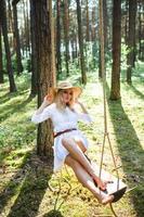 blond Jeune femme avec mince nu jambes dans blanc robe et paille chapeau séance sur une corde balançoire photo
