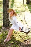 blond Jeune femme avec mince nu jambes dans blanc robe et paille chapeau séance sur une corde balançoire photo