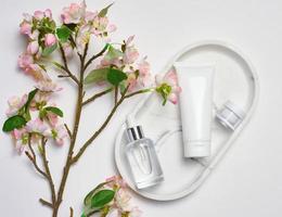 blanc Plastique baignoire, verre bouteille avec une compte-gouttes sur une marbre rester, et une bouquet de épanouissement Cerise sur une blanc arrière-plan, cosmétique récipient, Haut vue photo