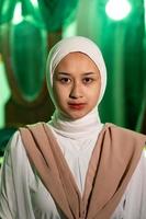 une musulman femme avec une blanc foulard et blanc vêtements est séance dans le milieu de une vert pièce sans pour autant maquillage photo