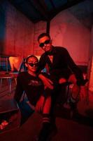 une couple de gay adolescents sont séance ensemble portant des lunettes de soleil et posant vilain dans une entrepôt avec une rouge lumière photo