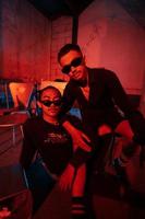 une couple de gay adolescents sont séance ensemble portant des lunettes de soleil et posant vilain dans une entrepôt avec une rouge lumière photo