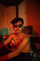 une gay homme dans des lunettes de soleil et denim un pantalon est assis relaxant dans une club à célébrer photo